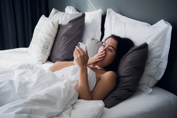 Mujer joven conmocionada mirando el teléfono móvil en la cama en casa — Foto de Stock