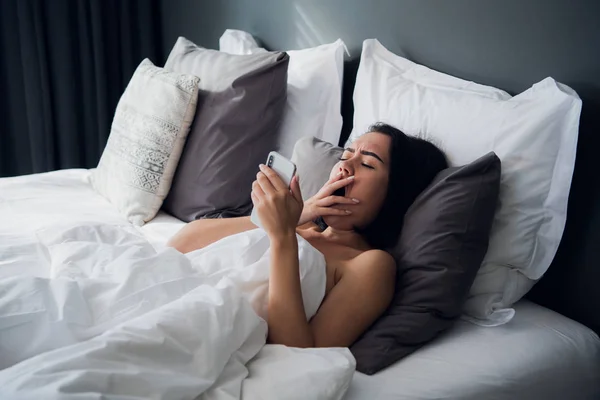 Foto de una mujer joven y bonita en el interior de su casa usando el chat del teléfono móvil. El bostezo yace en la cama . — Foto de Stock