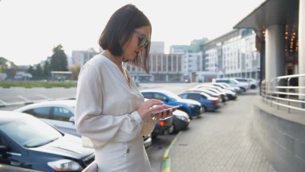 Mujer con blusa blanca enviando mensajes de texto en el teléfono inteligente caminando por la calle en un día soleado — Vídeos de Stock