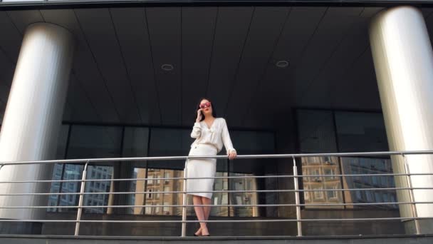 Hermosa mujer de negocios morena elegante con teléfono móvil hablando, sonriendo y riendo frente al edificio de rascacielos donde trabaja — Vídeos de Stock