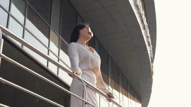 Mladý bělošský obchodní žena drží na zábradlí a shlíží na město při západu slunce. Brunetka má slunce a teplý vítr v létě v městě před kancelářská budova scyscraper ve zpomaleném záběru. — Stock video
