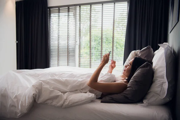 Chica morena está leyendo el mensaje de texto de su novio en el móvil, mientras está acostado en la cama. Alegre joven mujer cubierta por manta está viendo mensaje de vídeo en el teléfono celular antes de después de dormir . — Foto de Stock