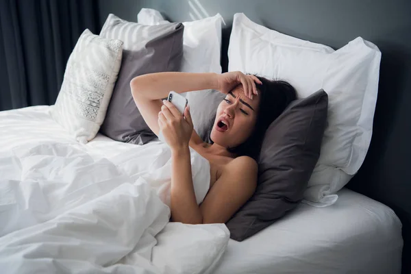 Imagen de una mujer frustrada de 20 años con cabello oscuro acostada en la cama sobre una almohada blanca después de dormir y mirando el teléfono inteligente móvil moderno con confusión — Foto de Stock