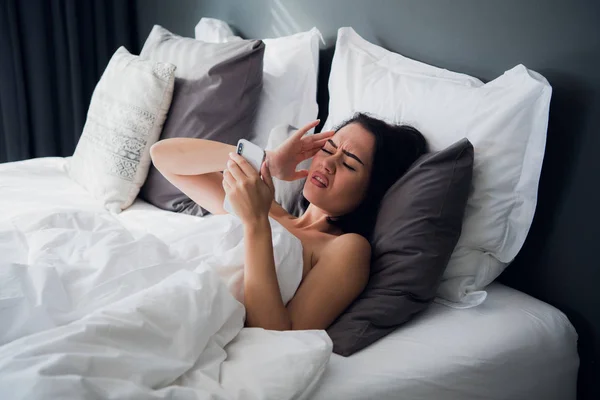 Portrait of a young brunette girl lying in a bed at home with a headache and back pain. Suffering from fever or flu and feeling pain at home. — Stock Photo, Image