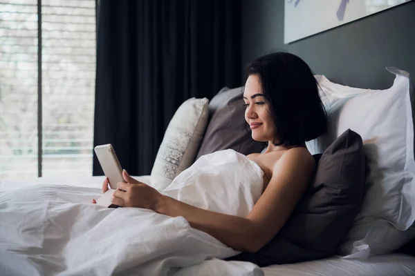 Retrato de la joven mujer caucásica feliz sentada en la cama, usando tableta digital y sonriendo — Foto de Stock