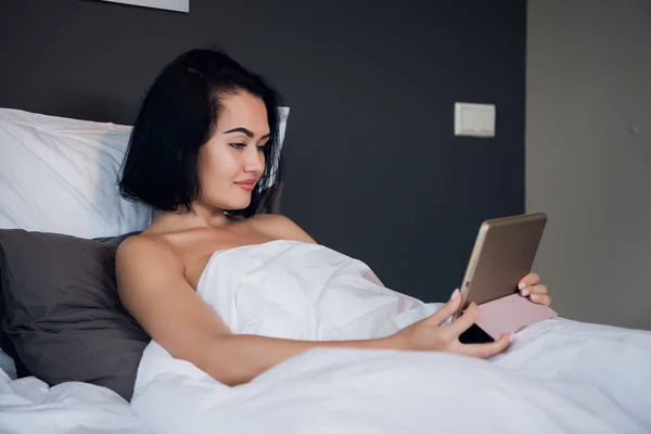 Retrato de la joven mujer caucásica feliz sentada en la cama, usando tableta digital y sonriendo — Foto de Stock