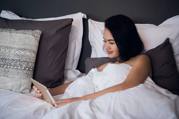 A smiling woman lies on the bed scrolling through her tablet.