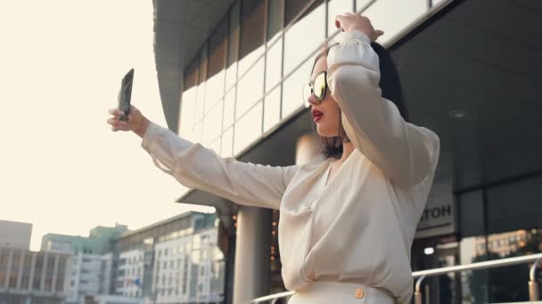 Fiatal gyönyörű barna üzletasszony mosolygó arcok így pózolt selfie fénykép használ smartphone kamera előtt office building, napnyugtakor a lassú mozgás a városban. — Stock videók