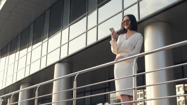 Slow-motion shot van gelukkige jonge die advocaat is praten over telefoon permanent op stad bouwen achtergrond, gelooid brunette in witte blouse bespreekt zaken met vrolijke glimlach buitenshuis. Concept: business — Stockvideo