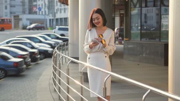 Mulher de negócios elegante bonita andando pela cidade conectada usando telefone inteligente. Senhora feliz vestindo blusa branca e saia mensagens ou redes sociais de navegação em seu telefone inteligente ao ar livre em lento — Vídeo de Stock