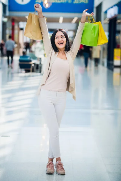 Egy sötét hajú nő visel tompa, gyengéd színekkel rendelkezik, színes, mintás bevásárló táskák sétál egy exkluzív shopping mall. — Stock Fotó