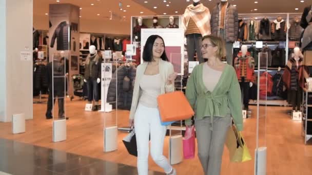 Glücklich lächelnde Frauen, die mit Tüten aus dem Kleiderladen kommen. Freundinnen gehen nach dem Einkauf vom Eingang eines Luxusgeschäfts in einem Einkaufszentrum. Zeitlupe. — Stockvideo