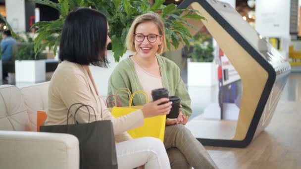 Dos mujeres jóvenes tomando café, hablando y riendo en el café en el centro comercial después de ir de compras. Gente, amistad, conversación y concepto de unión — Vídeo de stock