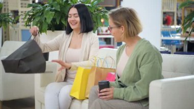 Kadın bir bluz alışveriş çantası gösterir dışarı onun arkadaşı alır, iki kadın gülümsüyor musun, arkadaş butikten bir kanepede oturan bir kıyafet satın tadını çıkarıyor, mutlu müşteri dostu konuşuyorsun