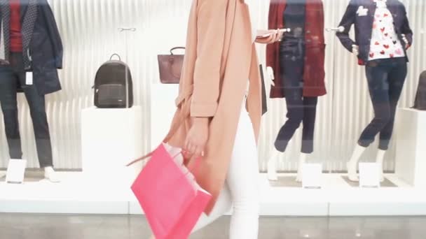 Mujer joven camina a lo largo de un escaparate con bolsas y teléfono en la mano en el centro comercial. Vista lateral dolly shot en cámara lenta . — Vídeos de Stock