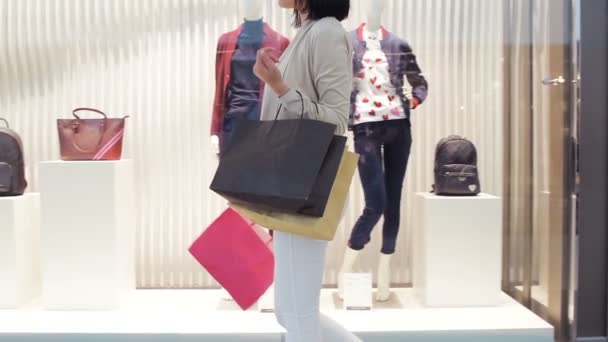 Young woman walks along a show window with bags and phone in her hand in the shopping mall. Side view dolly shot in slow motion. — Stock Video