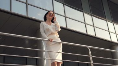 Gülümseme ve başarı. Genç kadın modern ofis binaları önünde telefonla konuşuyor. Ağır çekim.