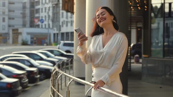 Aantrekkelijke jonge vrouw lopen in de stad. Het typen van een bericht op haar Smartphone. Vrouw wandelen, rust na het werk. Charmante glimlach. Officieel gekleed. Zonsondergang. Vervoer op de achtergrond — Stockvideo
