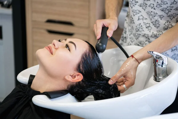 Cabeleireiro profissional irreconhecível lavar cabelo para seu cliente. Cabeleireiro bonito lavar o cabelo para sua senhora cliente no salão de cabeleireiro. Cliente sentado com os olhos fechados . — Fotografia de Stock