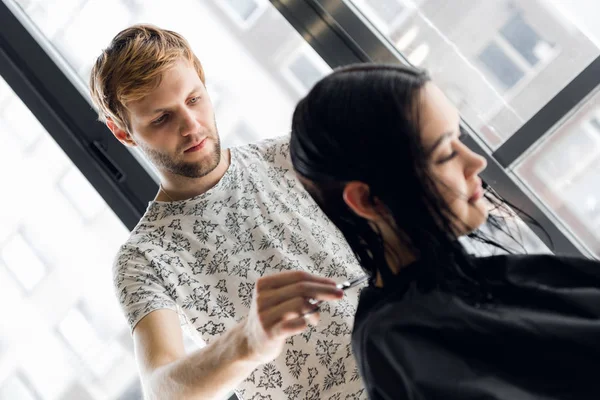 Cabeleireiro masculino sorrindo e conversando com um cliente enquanto faz um novo corte de cabelo para a jovem morena bonita — Fotografia de Stock