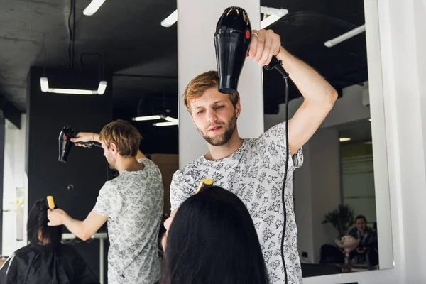 Чоловічий перукар посміхається і розмовляє з клієнтом, роблячи нову зачіску, щоб бути красивою молодою брюнеткою — стокове фото