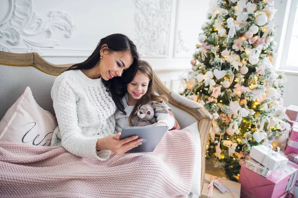 Mère avec fille sur le canapé en utilisant une tablette Happy Souriant jeune famille près décoré Nouvel An arbre de Noël petite fille et femme . — Photo