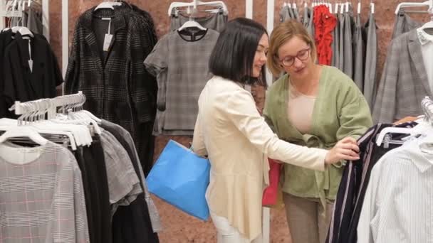 Mujer joven elige un vestido en una tienda de ropa y consulta con su amigo. Probándose vestidos en el espejo. Chica morena va a comprar un vestido y consulta con un amigo rubio . — Vídeo de stock