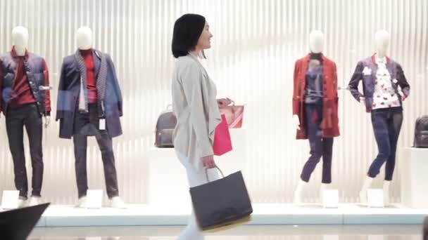Mujer joven camina a lo largo de un escaparate con bolsas y teléfono en la mano en el centro comercial. Vista lateral dolly shot en cámara lenta . — Vídeos de Stock