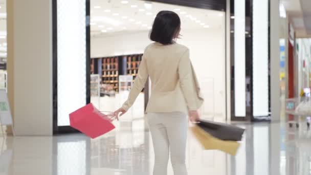 Mujer en jeans de colores suaves y chaqueta caminando y girando en el centro comercial mientras lleva bolsas de compras en ambas manos . — Vídeos de Stock