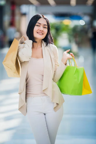 Een donkere harige vrouw dragen van gedempte, zachte kleuren houdt kleurrijke, patroon boodschappentassen loopt bij een exclusieve winkelcentrum. — Stockfoto