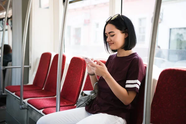 Joven adorable mujer alegre está sentado en el autobús usando el teléfono y sonriendo . — Foto de Stock
