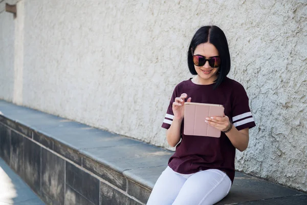 Emocionada joven feliz sosteniendo la tableta y apuntando con el dedo hacia arriba en el espacio de copia mientras está sentada aislada en la calle de la ciudad . — Foto de Stock