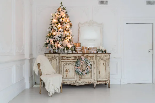 Vánoční stromek na dřevěné truhlici zásuvek prádelník v bílém interiéru, zdobené umělé květiny, girlandy a hračky — Stock fotografie