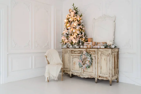Árbol de Navidad en la cómoda cómoda de madera en el interior blanco, decorado con flores artificiales, guirnaldas y juguetes — Foto de Stock