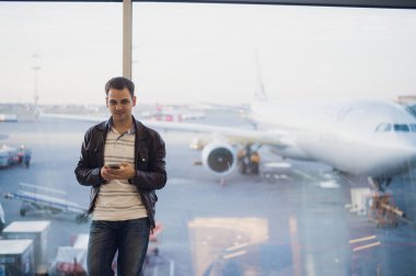 Gezgin Havaalanı terminal içinde. Cep telefonu kullanarak ve onun uçuş için bekleyen genç adam.