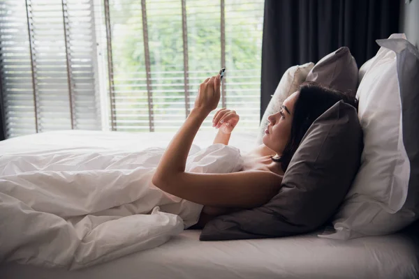 Feliz atractiva joven europea sonriendo ampliamente mientras lee el mensaje de texto de su novio usando el teléfono móvil en la cama, sin usar nada, cubriendo el cuerpo con una manta — Foto de Stock