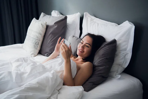 Chica morena está leyendo el mensaje de texto de su novio en el móvil, mientras está acostado en la cama. Alegre joven mujer cubierta por manta está viendo mensaje de vídeo en el teléfono celular antes de después de dormir . — Foto de Stock