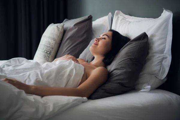 Retrato de una joven morena linda durmiendo en una cama en la sala de estar de luz. Su cabeza está sobre almohada gris y está cubierta con manta blanca. — Foto de Stock