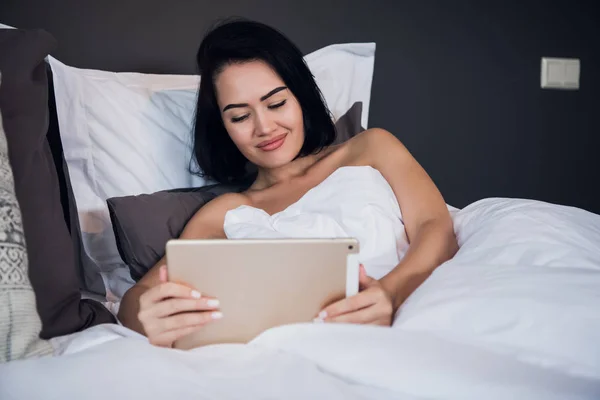 Una mujer sonriente yace en la cama desplazándose a través de su tableta. — Foto de Stock