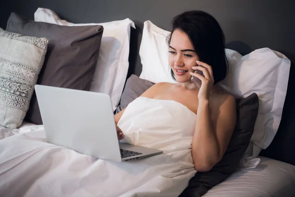 Sonriendo mujer joven relajada usando el teléfono móvil y el ordenador portátil en la cama en casa — Foto de Stock