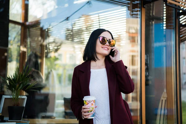 Al aire libre estilo de vida retrato de moda de la mujer joven y bonita hablando por teléfono. Sonriendo, caminando por la calle de la ciudad. Ir de compras. Usando un abrigo elegante, gafas de sol redondas — Foto de Stock