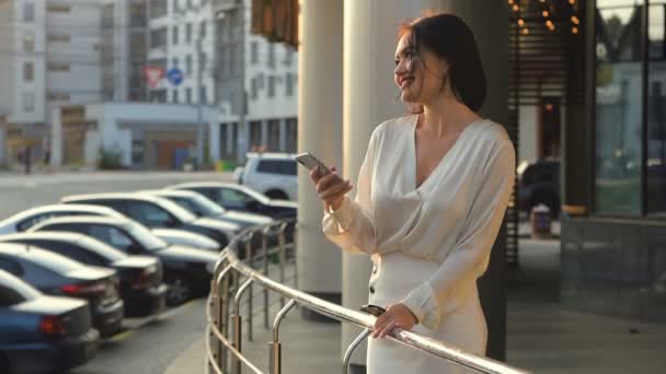 Mujer joven y feliz vistiendo camisa blanca usando teléfono móvil y sonrisa en la calle cerca de la carretera con coches en la ciudad europea en el día de verano — Vídeos de Stock