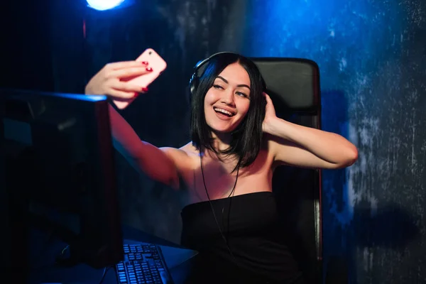 Alegre jugador streamer chica con auriculares y sentado cerca de su computadora PC tomando selfie en el teléfono durante el juego en línea —  Fotos de Stock