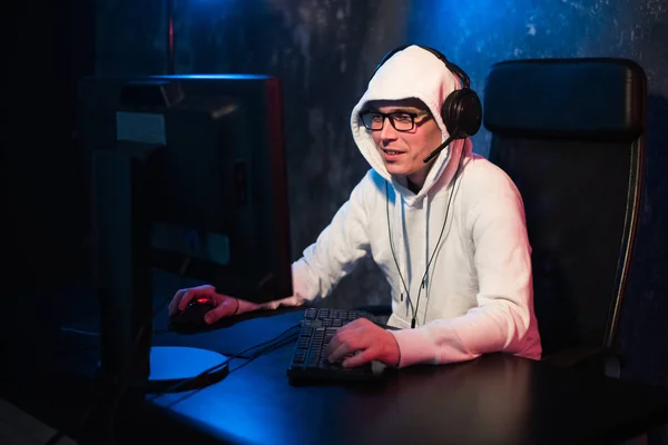 Porträt eines konzentrierten jungen Mannes mit Brille, Headset und Kapuze im dunklen Raum, der ein Online-Computerspiel spielt oder an einem Online-Esport-Turnier teilnimmt — Stockfoto