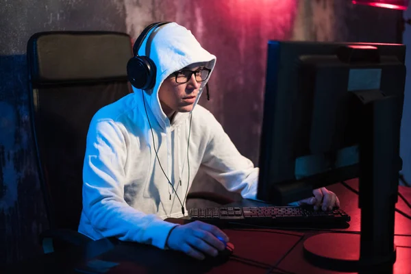 Retrato de un joven concentrado en gafas, auriculares y capucha en cuarto oscuro jugando juego de ordenador en línea o compitiendo en un torneo de esport en línea —  Fotos de Stock