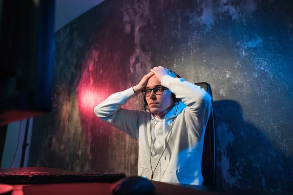 Divertido joven jugador con un auricular jugando videojuegos en una computadora y mira la pantalla con asombro. Retrato de un joven sorprendido usando una computadora por la noche —  Fotos de Stock