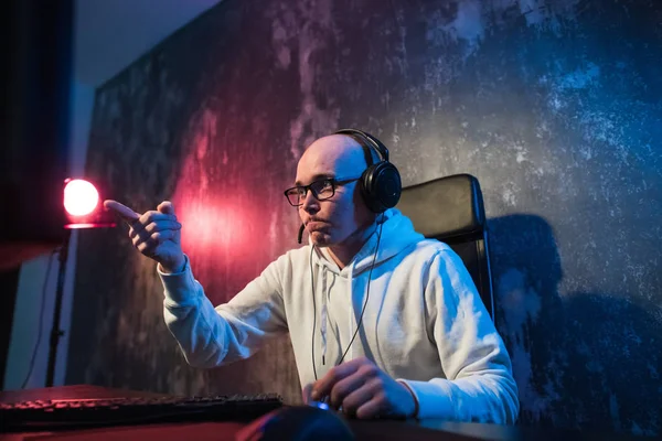 Divertido joven jugador con un auricular jugando videojuegos en una computadora y mira la pantalla con asombro. Retrato de un joven sorprendido usando una computadora por la noche —  Fotos de Stock