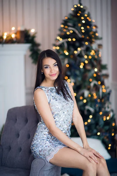 Різдво. Симпатична дівчина в блискучій сукні вдома сидить на кріслі проти каміна і ялинки — стокове фото