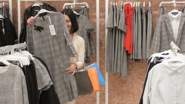 Δύο νεαρές γυναίκες περπατώντας μέσα από το εμπορικό κέντρο. Γυναίκα ψώνια. Νεαρή γυναίκα επιλέγει ένα sweather σε ένα κατάστημα ιματισμού και συνεννοείται με τον φίλο. Προσπαθώντας για φορέματα — Αρχείο Βίντεο