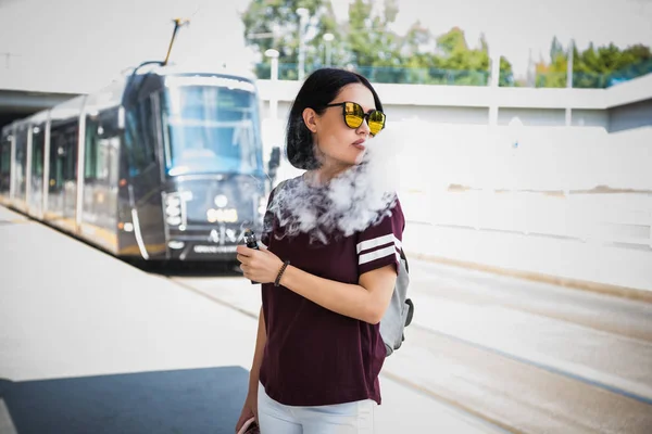 Bonita morena fumando un cigarrillo electrónico de pie en una calle exhalando una nube un humo de su boca mientras mira hacia un lado . — Foto de Stock